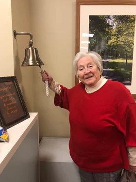 A companion in a red sweater rings a brass bell and smiles